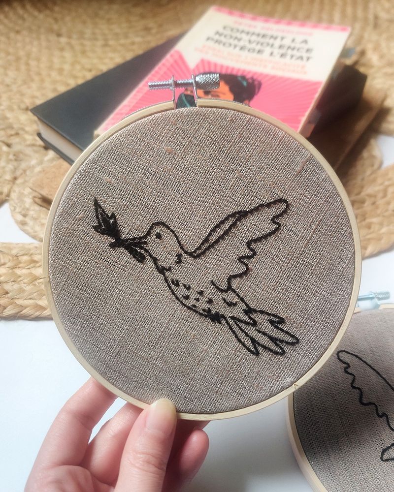 Photo d’une broderie d’un colibri avec une allumette dans le bec. Il est de profil et ses deux ailes sont vers l’arrière. Broderie dans un cercle en bambou.