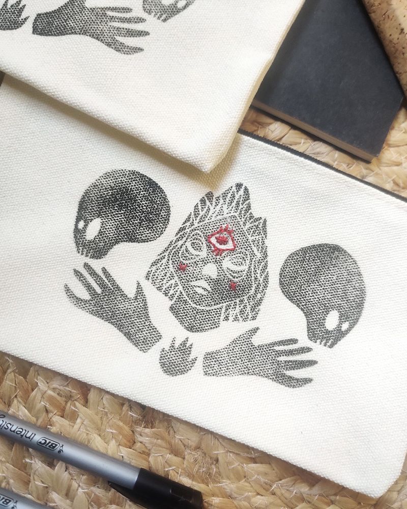 Photo d’une pochette en coton sur laquelle est gravée une illustration qui représente un portrait de femme entouré de deux crânes et souligné par une petite flamme, elle même entourée de deux mains tournées vers le haut, vers les crânes qui regardent vers l’extérieur. L’illustration est imprimée par la gravure, en noir, et un troisième oeil a été brodée au fil rouge sur le front du portrait.