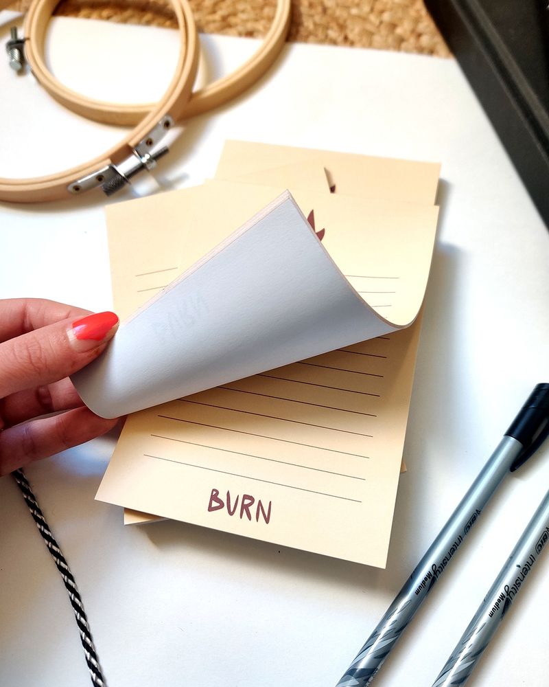 Photo d’un bloc notes au papier jaune très clair. Il y a une flamme au dessus des lignes et le mot "burn" en dessous. Sur cette photo, une mains soulève la première feuille du bloc notes.