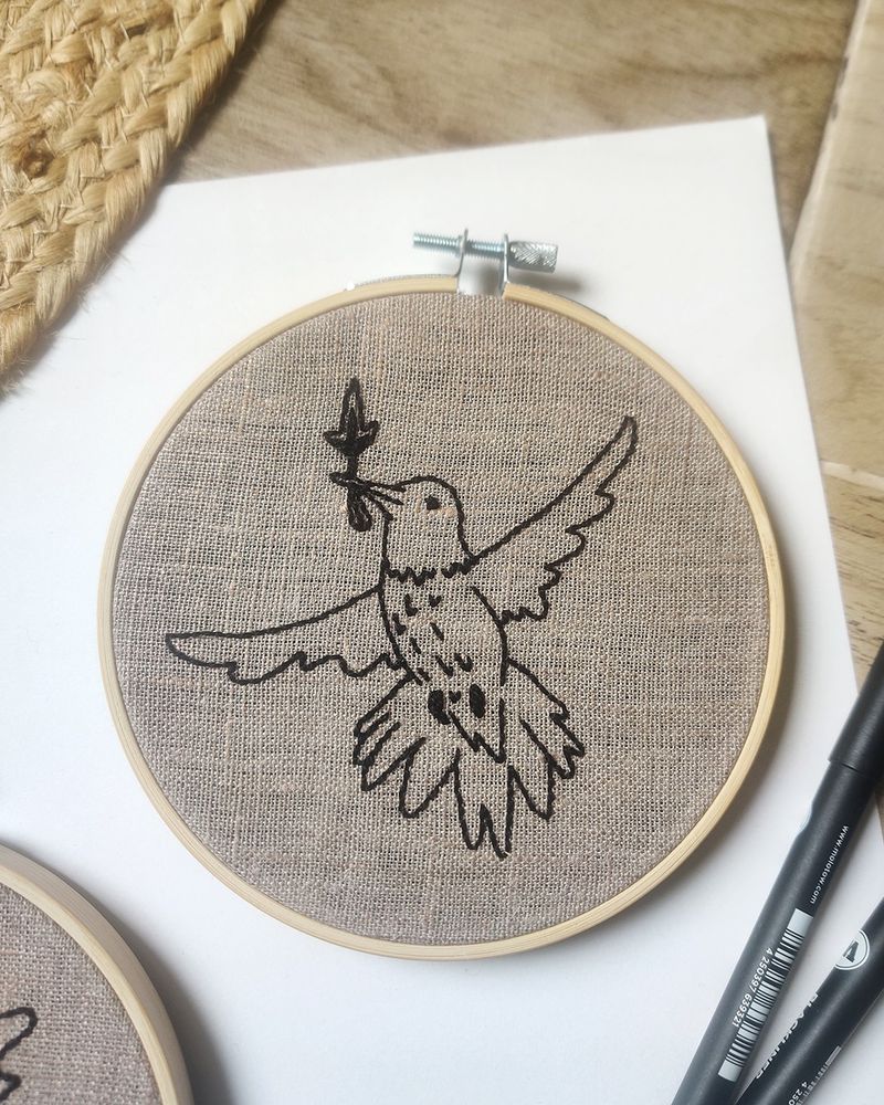 Photo d’une broderie d’un colibri avec une allumette dans le bec. Ses ailes sont déployés autour de lui et son regard va vers le haut. Broderie dans un cercle en bambou.