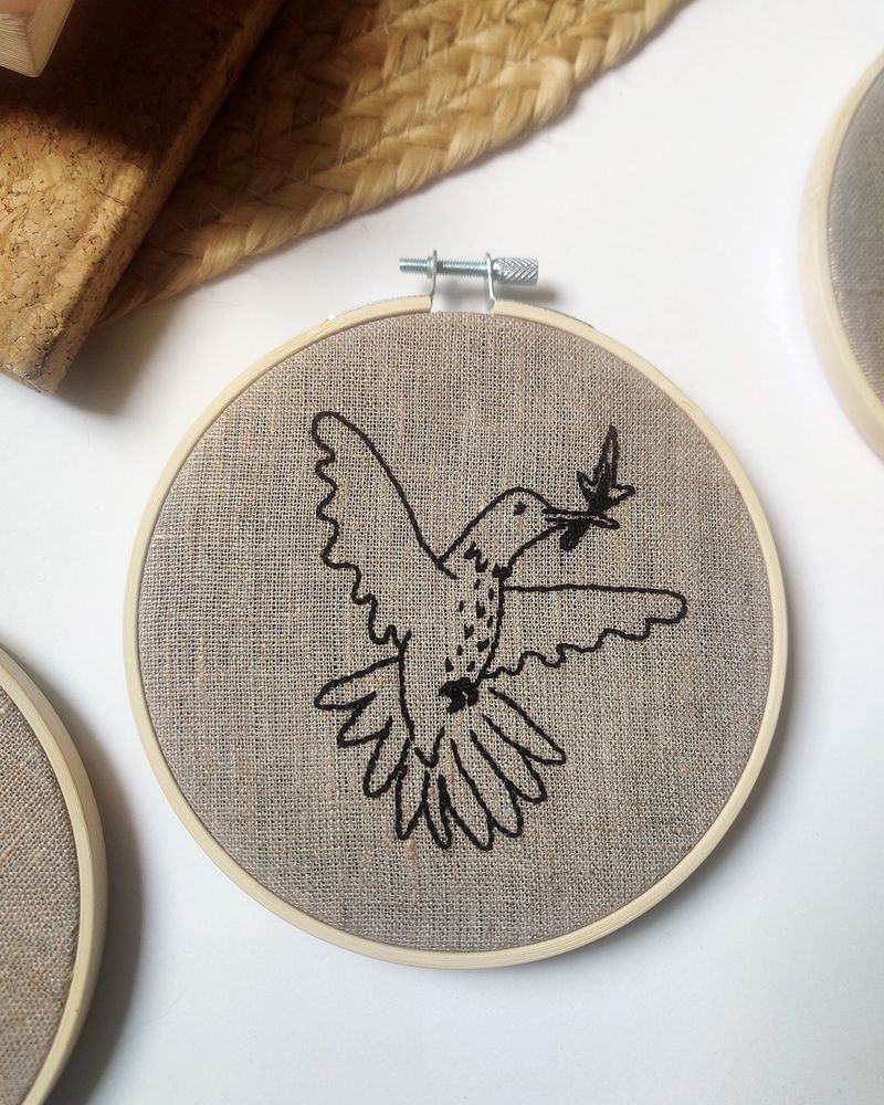 Photo d’une broderie d’un colibri avec une allumette dans le bec. Ses ailes sont déployés autour de lui et il regarde droit devant. Broderie dans un cercle en bambou.