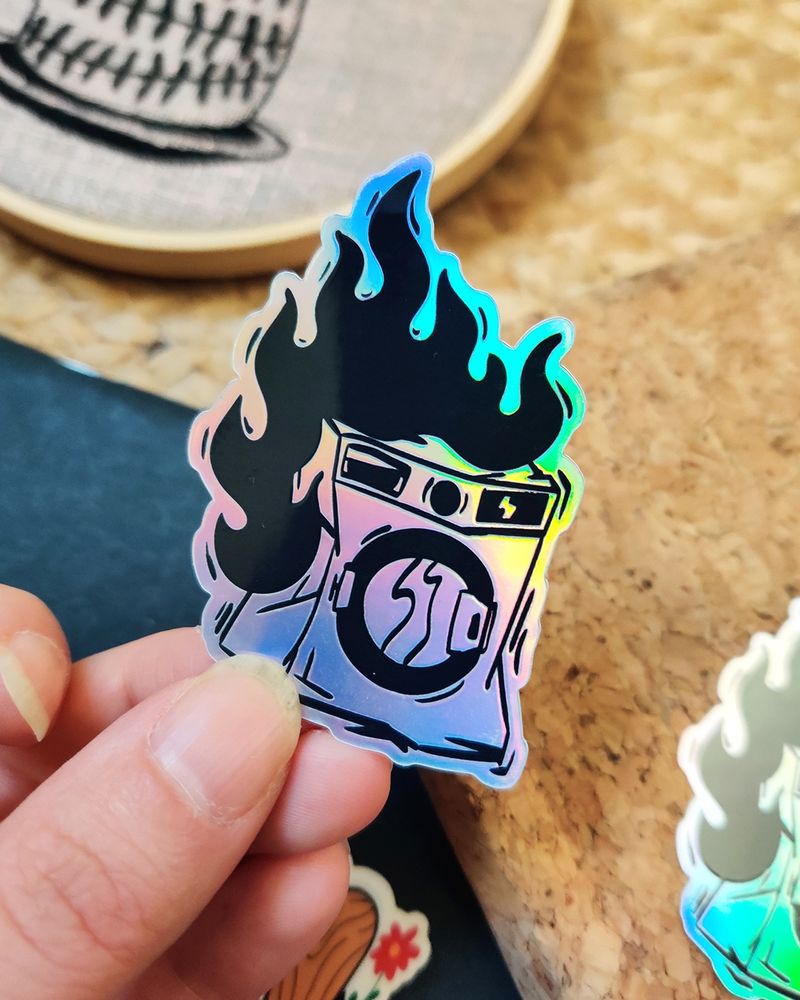 Stickers holographique qui représente une machine à laver toute cabossée et en flammes. Le stickers est tenu dans une main.