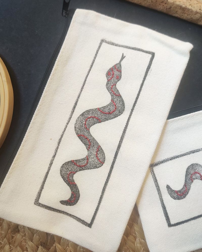 Photo d’une pochette en coton sur laquelle a été imprimée via la gravure la silhouette d’un serpent qui est entouré d’un cadre. Des motifs de cercles ont été ajoutés sur le serpent avec des broderies au fil rouge.