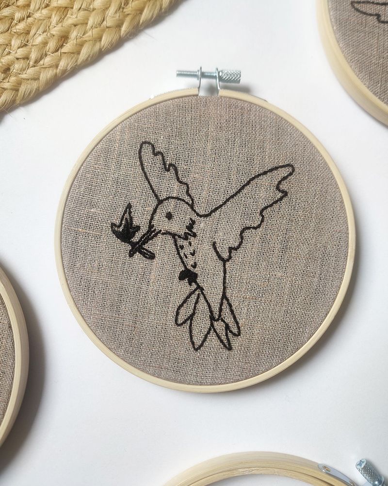 Photo d'une broderie d’un colibri avec une allumette dans le bec. Il regarde vers le bas et ses deux ailes sont vers l’arrière. La broderie est dans un cercle en bambou.