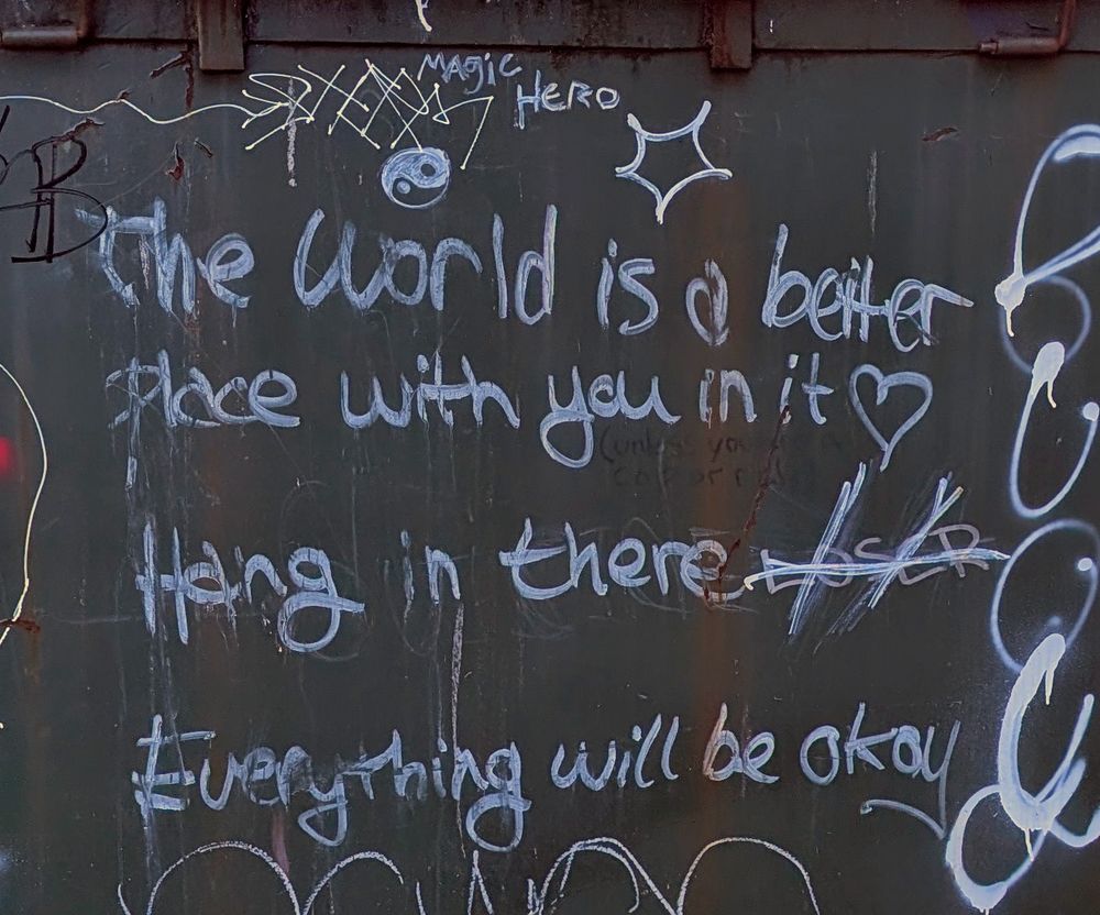 A dumpster with scrawled text that reads, "The world is a better place with you in it, hang in there, everything will be okay"