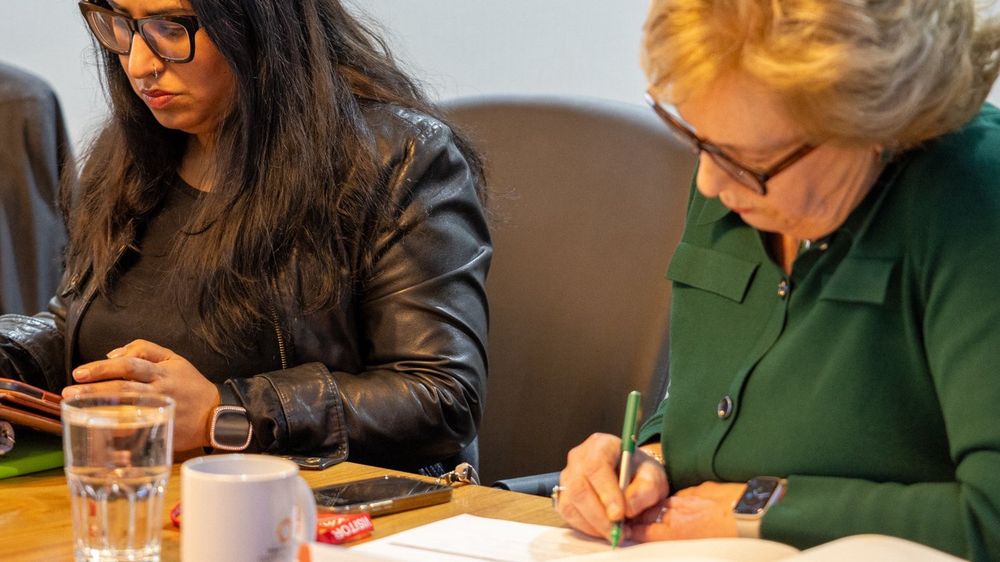 Photograph of two women sat down at a table writing and working on an i-Pad. 