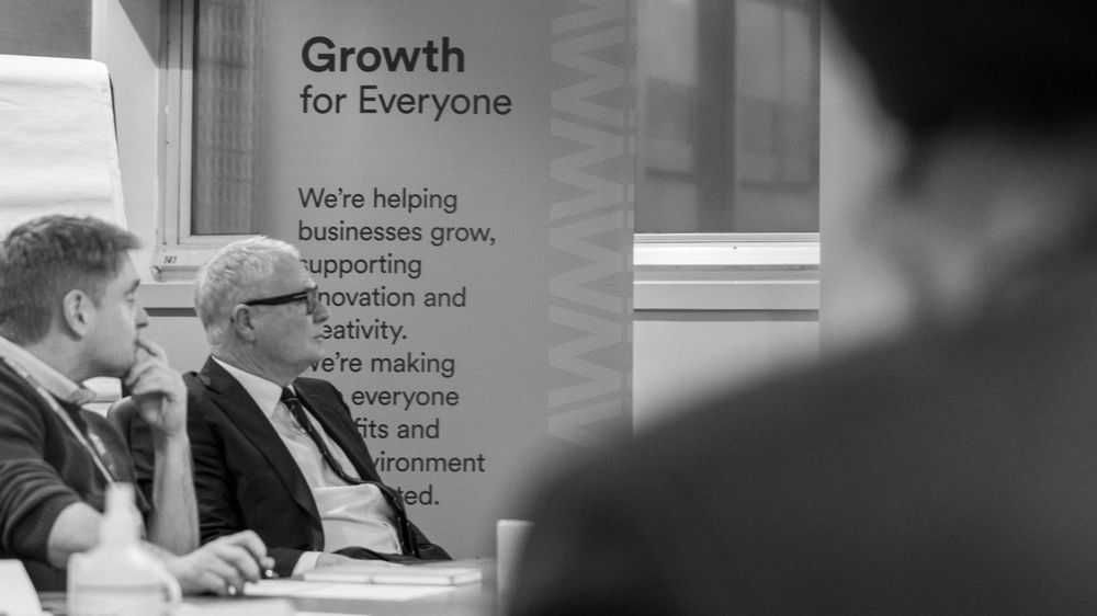 Photograph of Mayor Richard Parker sat down next to a man in a meeting with a pull-up banner that reads Growth for Everyone.