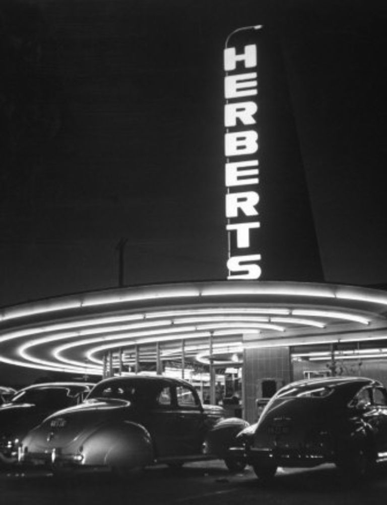 A black and white 1945 photo of Herbert’s drive-in restaurant on the southeast corner of Beverly Blvd and Fairfax Ave. The HERBERTS blade sign, the shiny pre-war cars that people were still driving because automobile production was halted for the duration of the war. There are concentric circles of neon on the underside of the overhang.