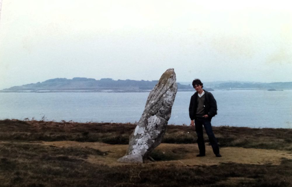The person posting is standing next to an inclined standing stone with the sea and islands behind.