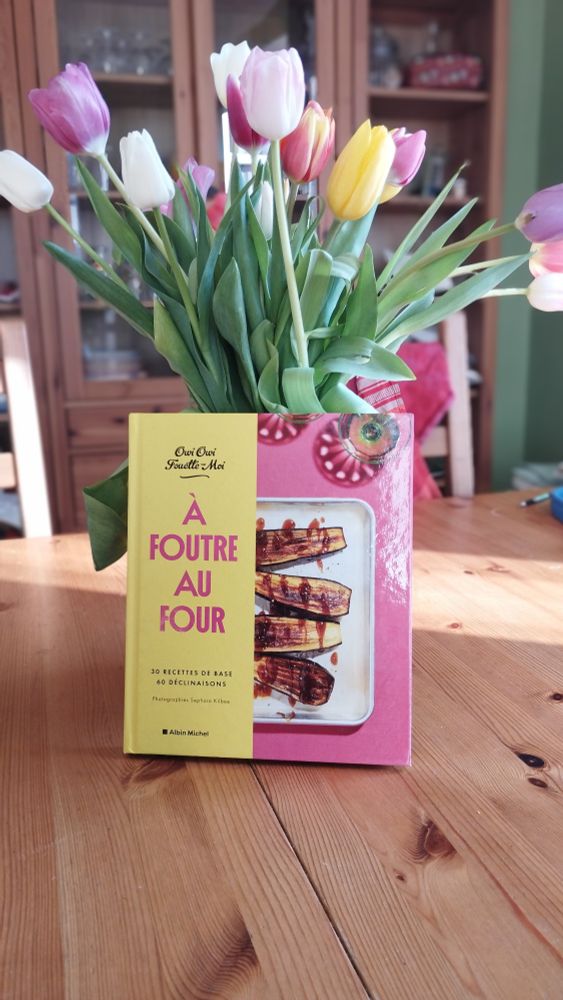 Livre de cuisine intitulé "à foutre au four", rédigé parOwi Owi fouette moi, adossé sur un joli bouquet de tulipes multicolore, le tout sur une table en bois 