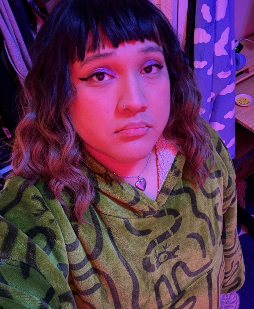 Selfie of Ray looking sad. She is wearing purple eyeshadow, winged eyeliner, a heart locket and oversized green sweater blanket. 