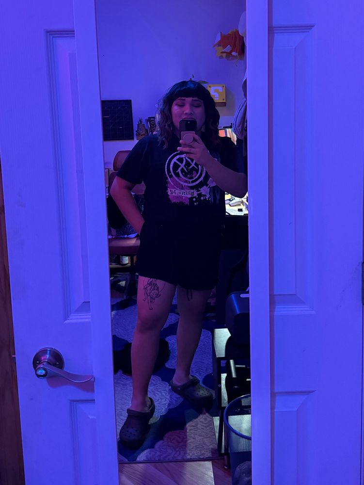 Mirror selfie of Ray. She is wearing her everyday wig (a curly bob), a blink 182 shirt and has short shorts that reveal her princess mononoke thigh tattoo. 