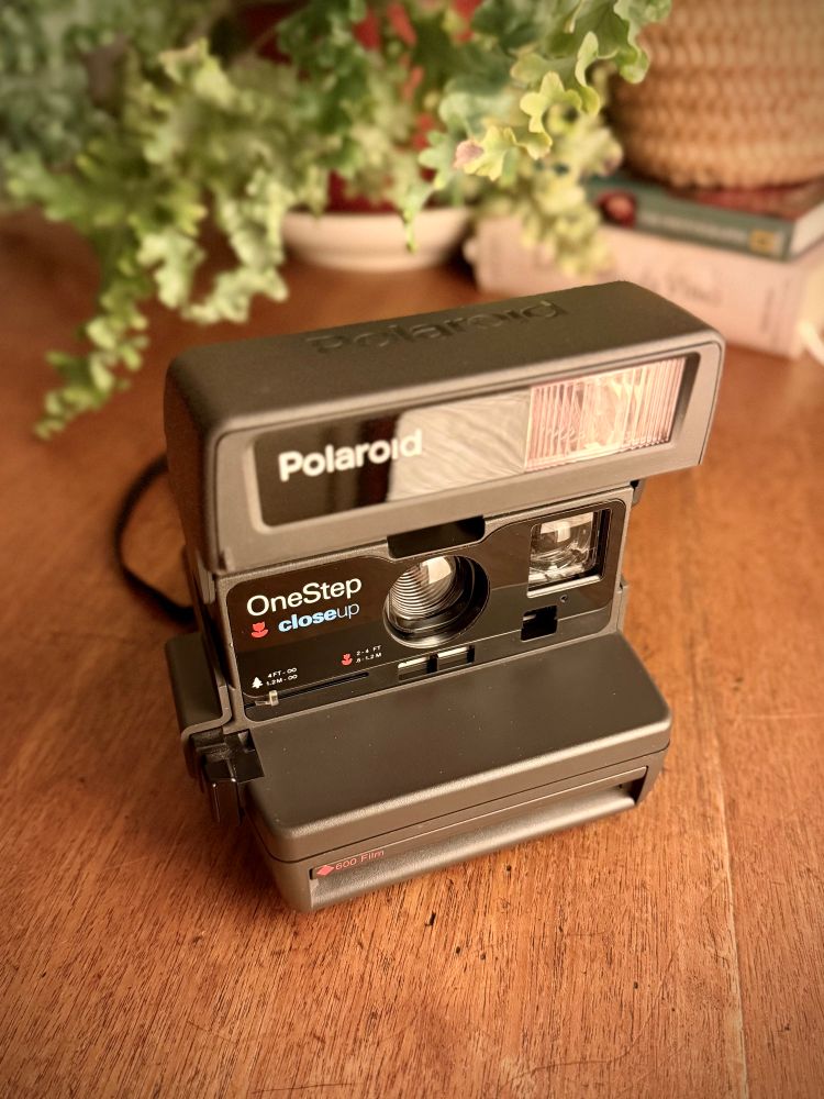 The Polaroid camera, dark grey with large flip up flash, with the words “Polaroid OneStep close up” on the front. The camera is sat on a wooden table in front of a couple of plants in pots. This is an iPhone snap for a product shot and has been toned and little and had vignette added. 