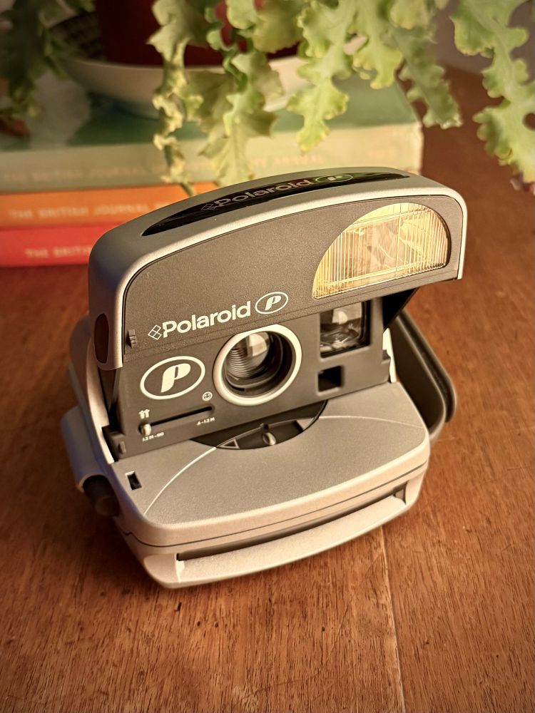 The Polaroid camera with a front flipped up, sat on a wooden table in front of some photography books. This is intended as a product shot and taken on an iPhone.