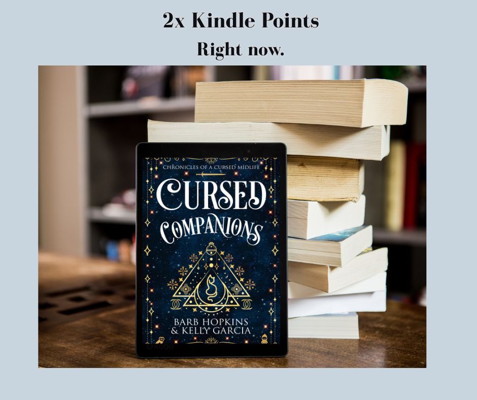 Ereader with Cursed Companions on it standing in front of stack of non-descript books on a wooden table. 