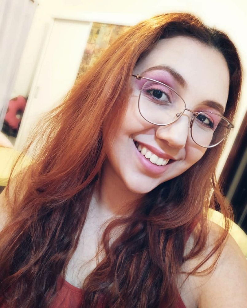 Selfie minha. Sorrio, o rosto inclinado e com óculos de armação fina. Meu cabelo está ruivo laranja, com a raiz mais lisa e ondulando no comprimento. O cabelo é tão comprido que não dá pra ver as pontas na foto. Gosto dessa selfie, estou cheia de sombra cor de rosa nos olhos. A foto é do começo de 2021