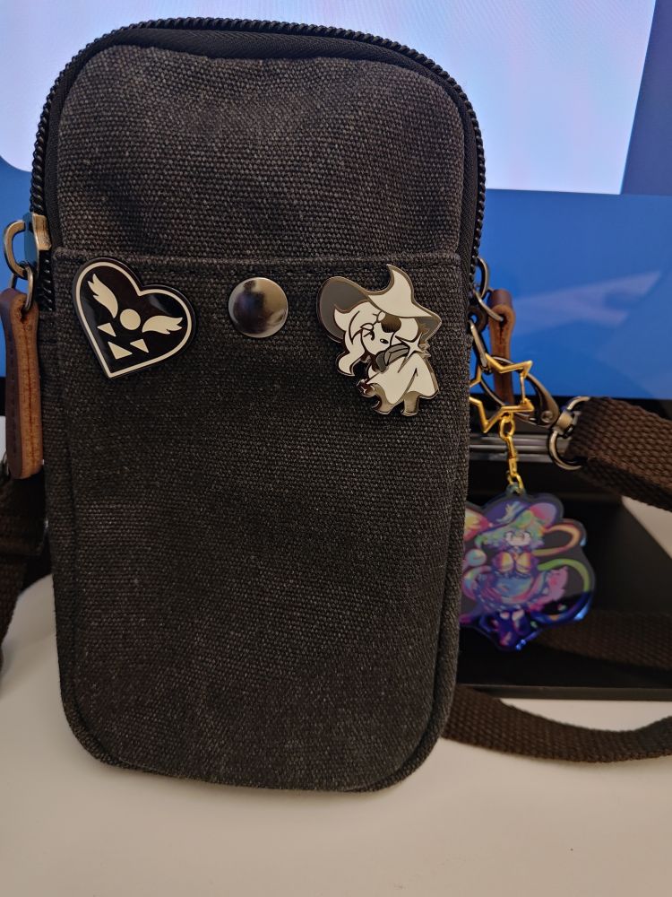 a small grey canvas bag. there is a deltarune pin and a siffrin isat pin on the front, and a koishi komeiji keychain attached to the side.