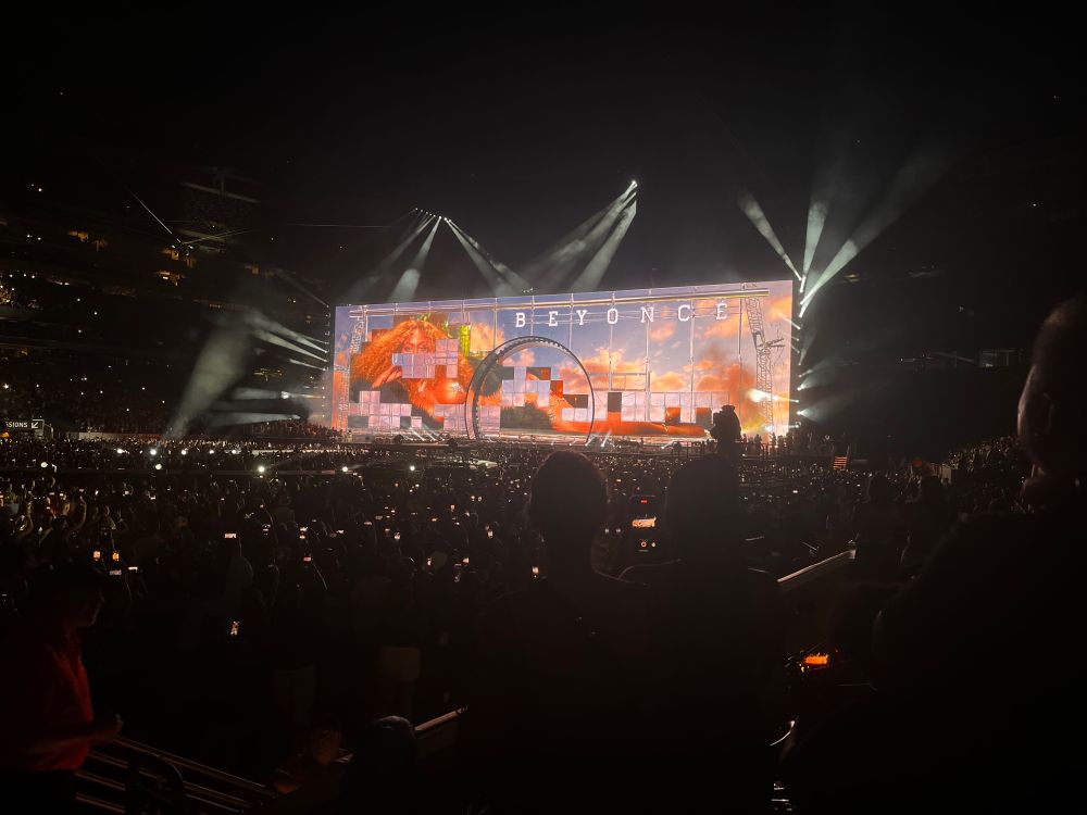On a giant horizontal screen in a stadium, an image of Beyonce lying on her side is partially visible, small sections hidden by squares in the shape of the period table of elements with a background of a sky at sunset