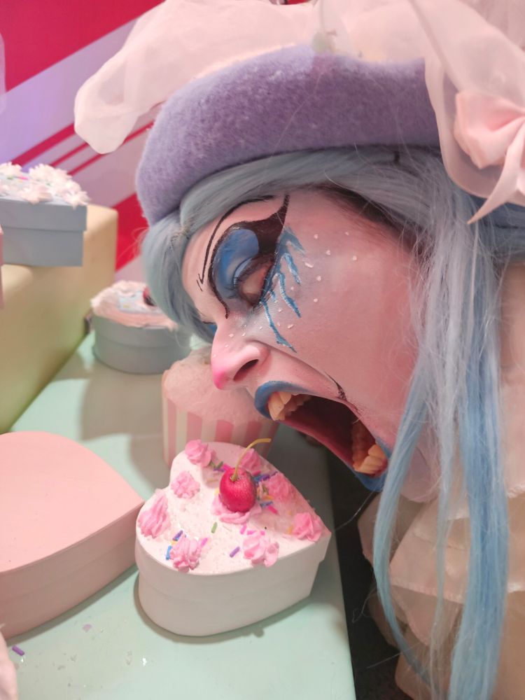 Humanette Honks, a hot clown, leans over a small heart-shaped prop cake, her mouth wide open, teeth bared as if to take a bite. She stares at the cake with a humorous level of intensity. Her look is winter-themed, all white and cool blue, with a flushed pink nose. Little icicles are drawn in eyeliner, dripping down her cheeks. She looks cute, silly, and hungry!