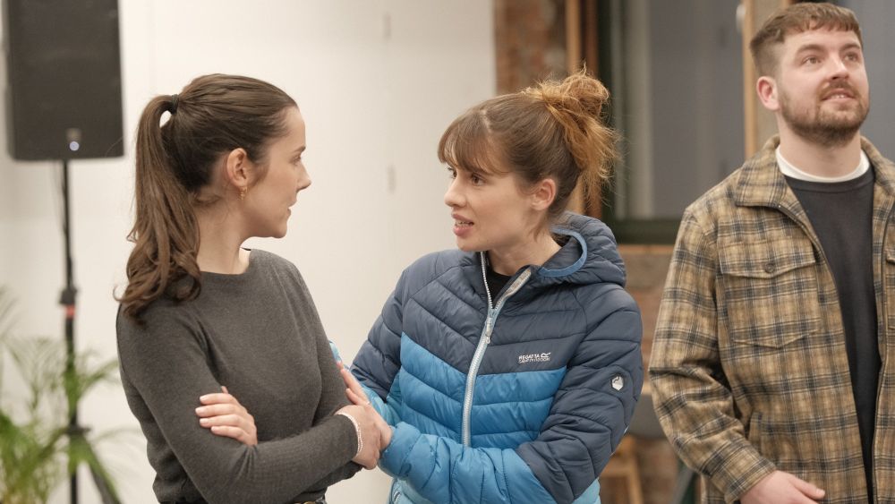 Actors Erin Elkin and Alex Paton stand together looking at each other. Erin is touching Alex's arm in a reassuring way. Jordan Monks stands in the background gazing upwards.