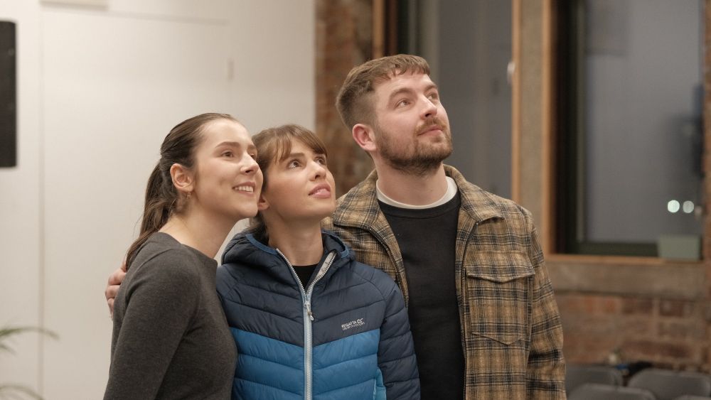 Actors Alex Paton, Erin Elkin and Jordan Monks stand huddled together looking upwards and smiling