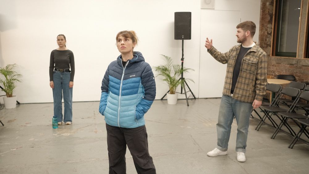 Actor Erin Elkin stands gazing upwards, while actor Jordan Monks stands  behind her with his arm outstretched. In the background actor Alex Paton stands still staring straight ahead.