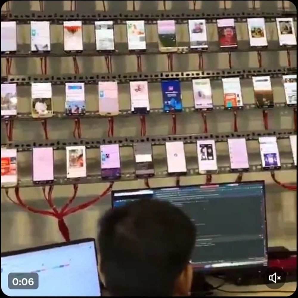 Bot- bzw. Trollfarm. An einer Wand sind in mehreren Reihen unzählige Handys angeschlossen. Ein Mann sitzt davor und betreut sie.