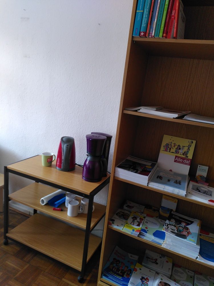 Ein Regal mit Infomaterial zu Kinderrechten. Daneben ein kleineres Regal mit Wasserkocher, Kaffeemaschine und Tassen.
