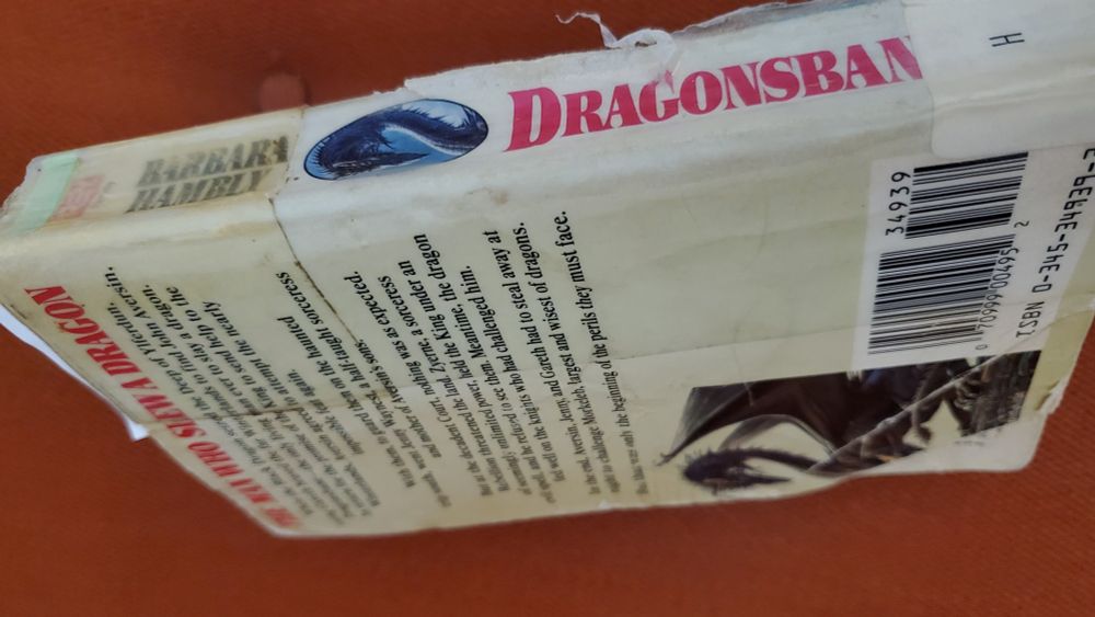 A battered copy of Barbara Hambly's Dragonsbane, propped on end so spine and back cover are in view, with a plastic library H marker wrapped around the spine over the E in Dragonsbane.