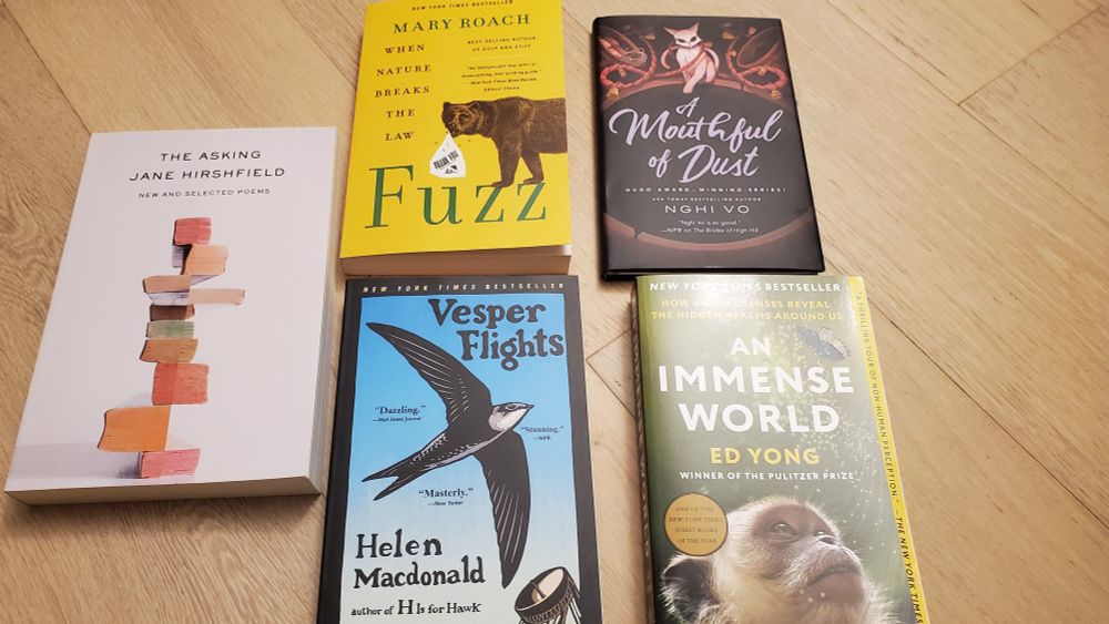 A picture showing the covers of 5 books: The Asking by poet Jane Hirshfield; Vesper Flights, an essay collection by Helen Macdonald; Fuzz, nonfiction by Mary Roach; A Mouthful of Dust, the newest novella in the Singing Hills cycle by Nghi Vo; and An Immense World, nonfiction by Ed Yong.