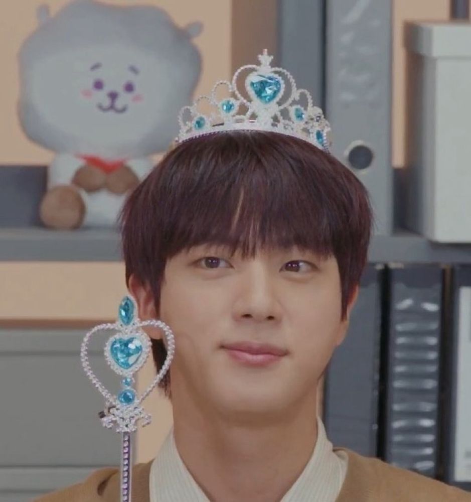 Close up of Jin of BTS wearing a silver plastic tiara with blue gems and holding a silver plastic wand that's shaped into a heart with blue gems. He is wearing a light brown cardigan and is sitting in front of a cabinet full of binders and a BT21 RJ plushie. He is giving a small smile towards something on his right, behind the camera. 
