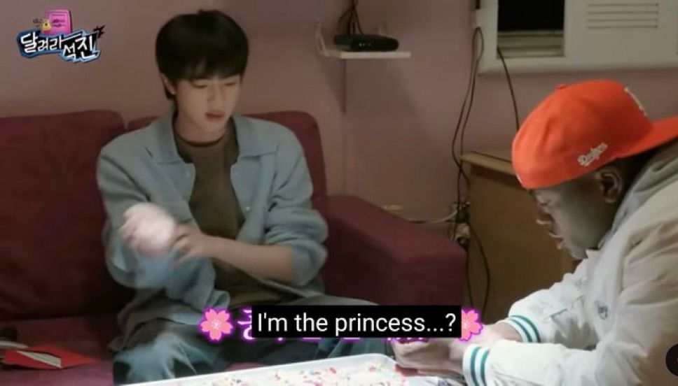 Screenshot of Jin of BTS sitting on a red couch, wearing a blue button up that's completely open and a green shirt underneath. His friend Jonathan sits across from him wearing a red-orange hat and is leaning across the low table they are both sitting around and looking at. Caption from video days "I'm the princess...?"