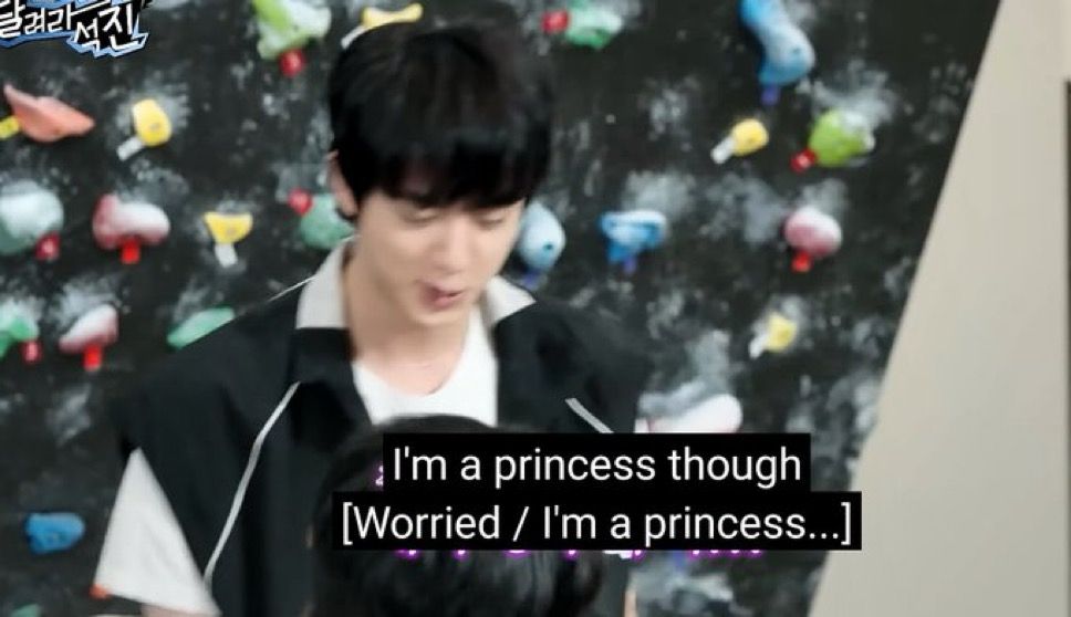 Screenshot of Jin of BTS standing in front of a rock climbing wall, looking down and talking to his show's guest. Said guest's head is just barely seen above the bottom half of the captured video still. Caption from video says "I'm a princess though [Worried/I'm a princess...]