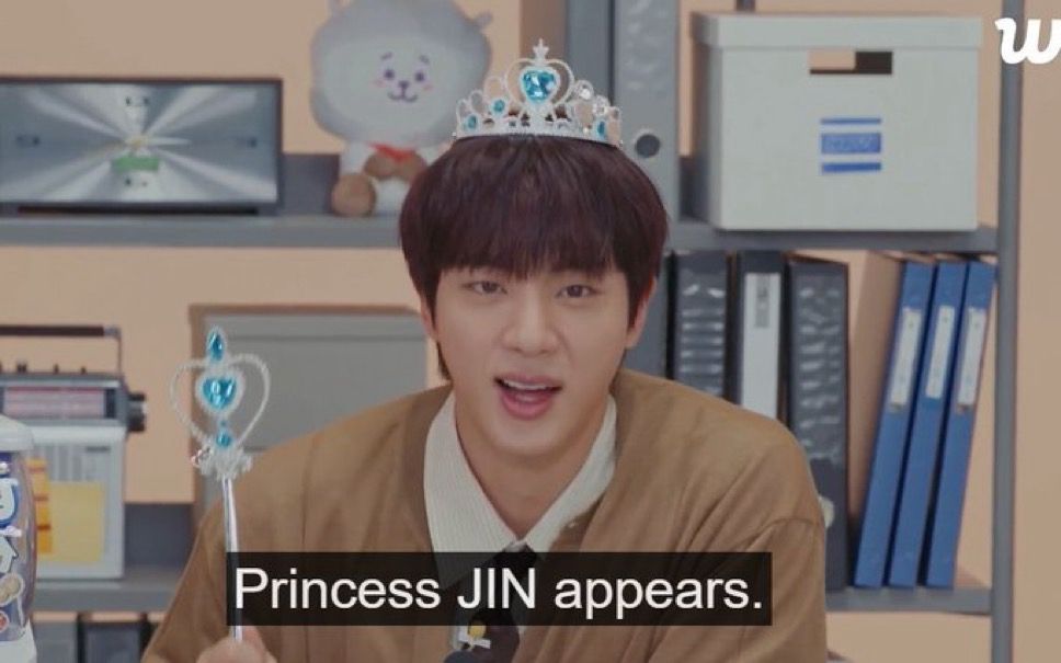 Screenshot of Jin of BTS wearing a silver plastic tiara with blue gems and holding a silver plastic wand that's shaped into a heart with blue gems. He is wearing a light brown cardigan and is sitting in front of a cabinet full of binders, a radio, and a BT21 RJ plushie. Caption from video says 'Princess JIN appears."
