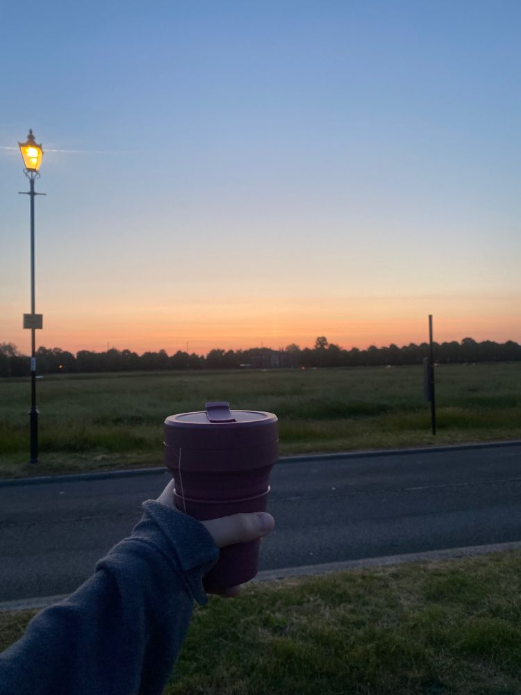 A sunrise sky with my arm out holding a burgundy coffee cup in the foreground
