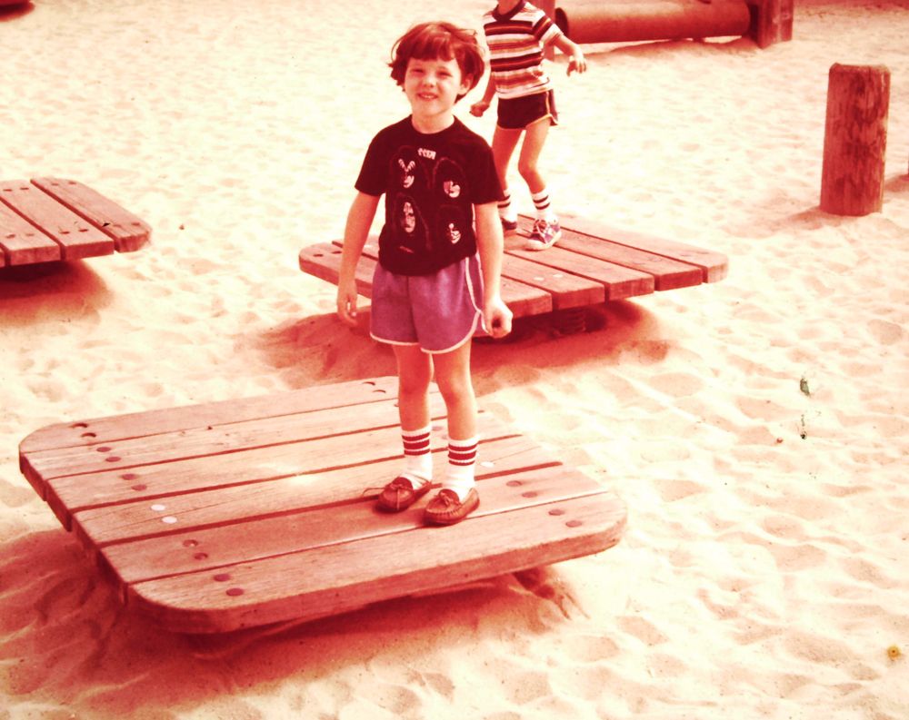 Five year old boy in a Kiss t-shirt and moccasins, squinting into the sunlight at a playground circa 1980. The picture has a sepia tone feel to it, as it’s a scan from an old Polaroid.