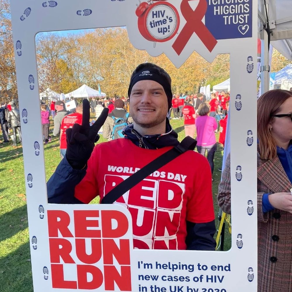 Man in a red "RED RUN LDN" shirt and beanie, smiling and making a peace sign, holds a frame promoting World AIDS Day and ending HIV in the UK by 2030.