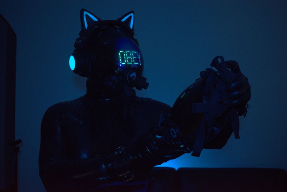 Drone 8012 stands, holding a MSA millennium, inspecting the gasmask in it's hands very closely. It is dressed in all black latex with a black corset with blue highlights. It is wearing it's MSA millennium with it's signature blue visor with the word OBEY written on it.