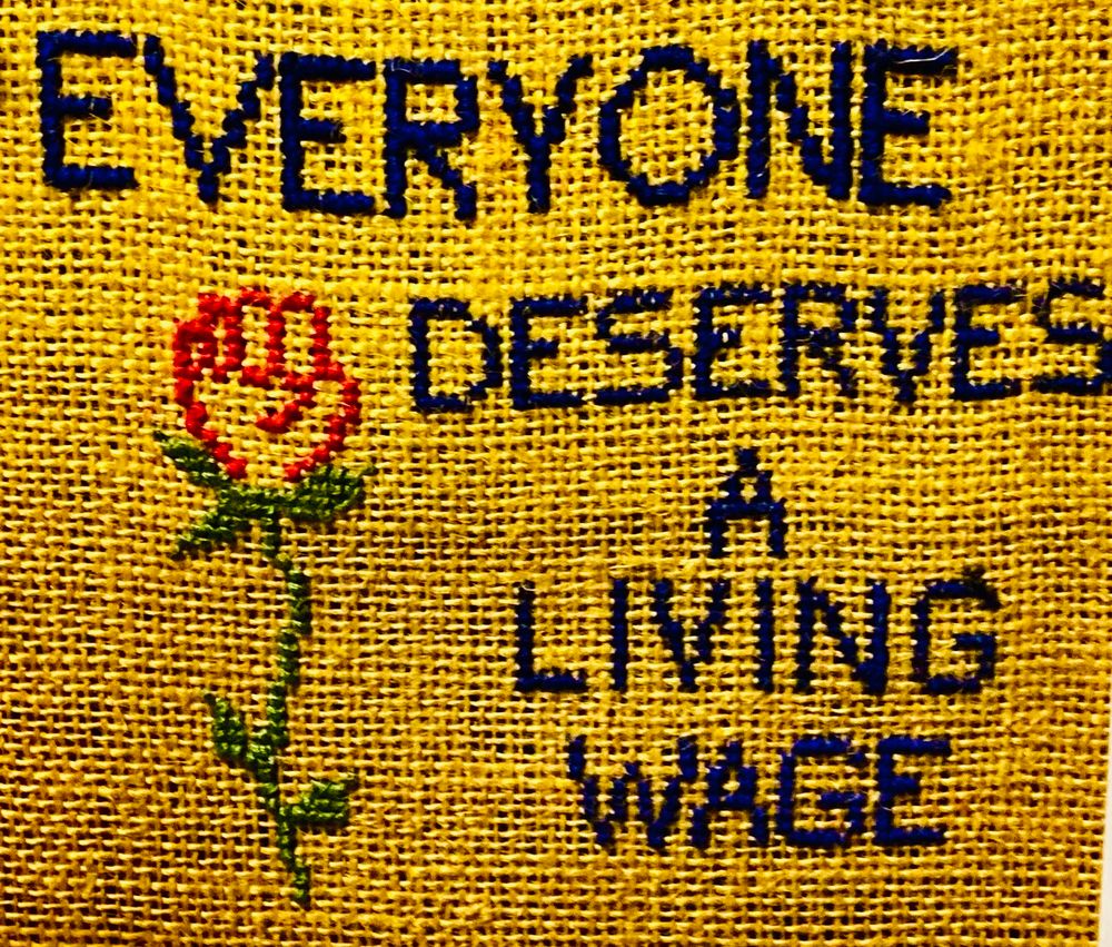 On yellow hessian, embroidered black text in capitals reads ‘everyone deserves a living wage’. There’s an embroidered red rose on a green stem to the left of the text. The red bloom is in the shape of a fist.