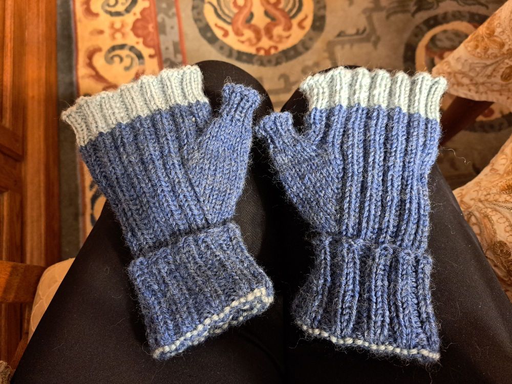 A pair of knitted fingerless mitts with a folded up cuff. The line of the fold and a thick stripe at the top edge of the fingers is light green while the main color is heathered blue. The main stitch pattern is k2p2 ribbing, the thumb gusset is stockinette and the last inch or so of the thumbs is k1p1 ribbing.