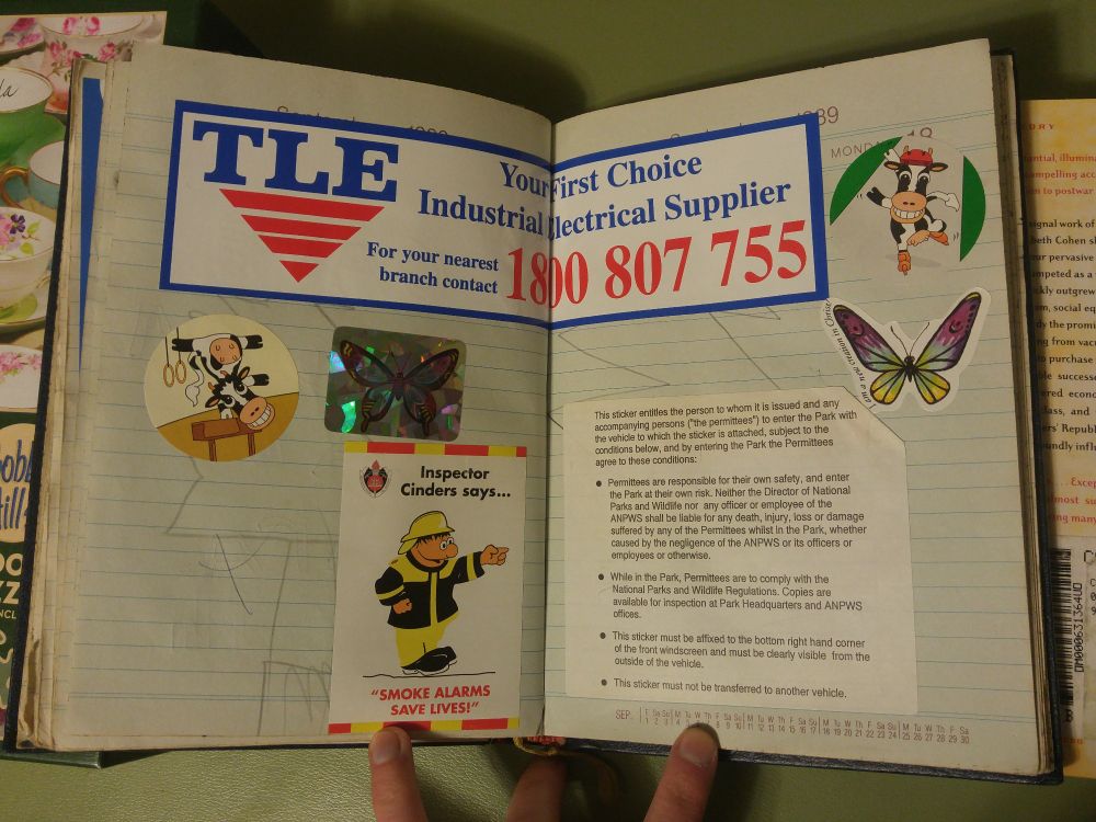 A spread of an old day planner, with a bunch of random stickers. Two butterflies. A bumper sticker tor TLE Your First Choice Industrial Supplier for your nearest branch contact 1800 807 755, two circular stickers of cartoon cows (one rollerblading, the other doing gymnastic vaulting, a cartoon firefighter "Inspector Cinders says smoke alarms save lives". A large square of text that's a national parks pass.