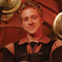 Hoban "Wash" Washburne, the pilot from Firefly and Serenity, portrayed by Alan Tudyk. Image is a close up of a promotional still featuring Tudyk in costume looking down the barrel of the camera with a gentle smile.