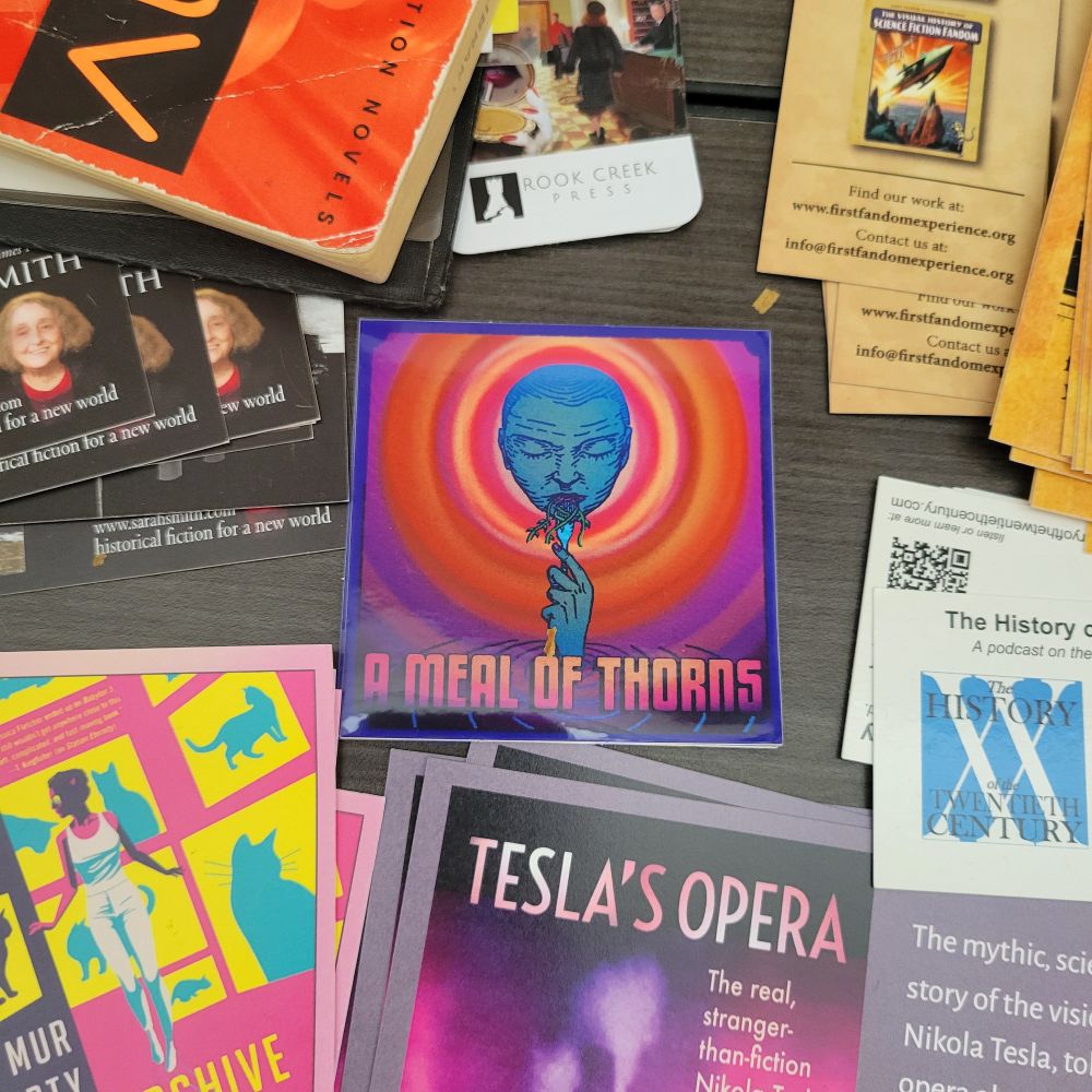 A table with bookmarks, flyers, and free books, with one square sticker with the Meal of Thorns logo.