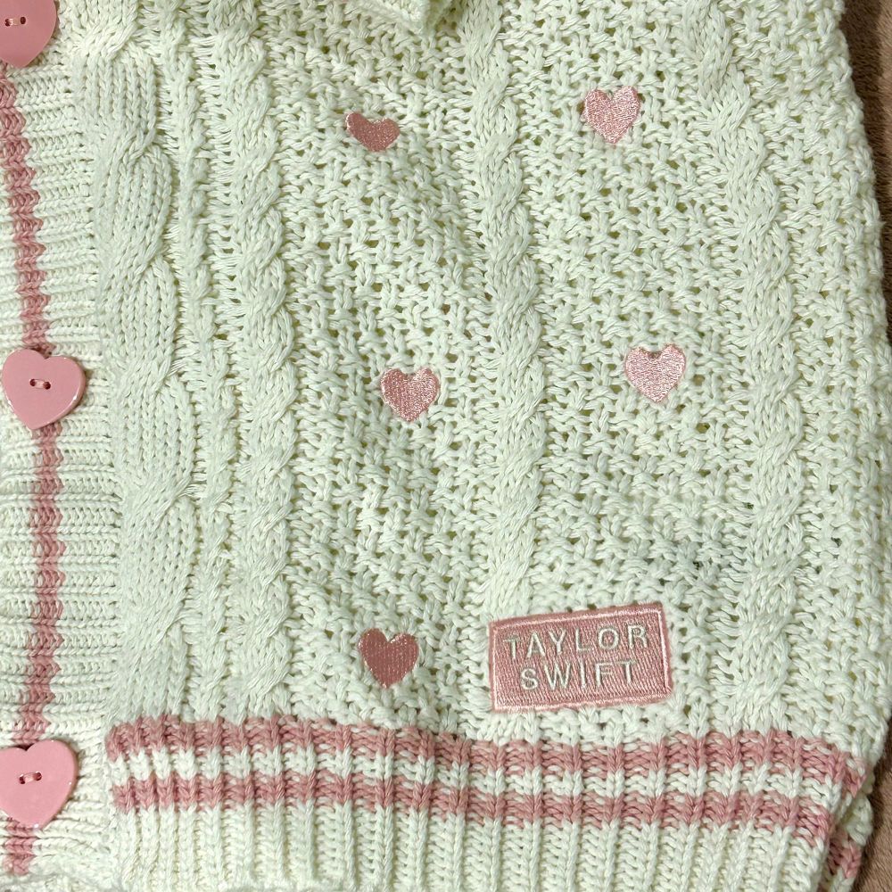 close up photo of the bottom corner of Taylor Swift’s lover cardigan, you can see the white knit cardigan with two baby pink stripes along the hem, a pink patch which says Taylor Swift, little pink hearts on the actual cardigan and pink heart shaped buttons 