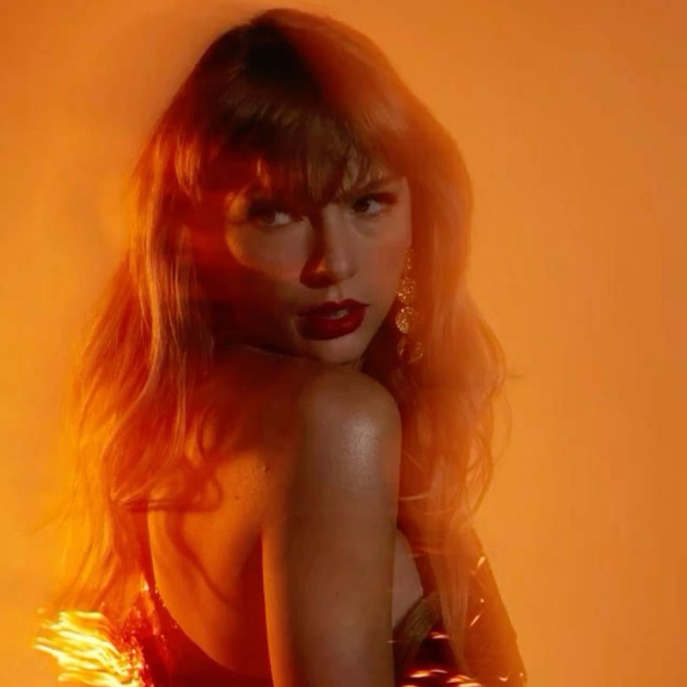 Taylor Swift stood in front of an orange background, she’s looking over her shoulder and there’s a bit of a blur behind her 