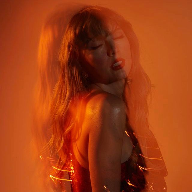 Taylor Swift against an orange background she’s stood on the side with her eyes closed and it’s blurry