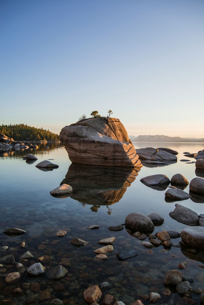 This photo by Ethan Dow shows a large granite boulder known as Bonsai Rock surrounded by clear water and smaller rocks at Lake Tahoe during a calm sunset, with pine trees and mountains in the background.