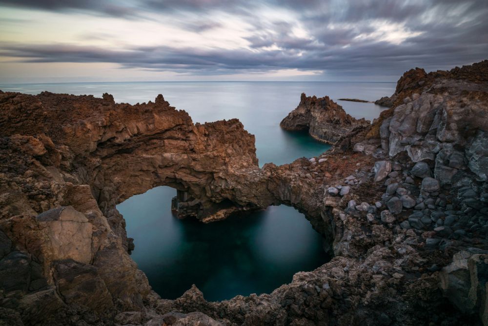 This photo by Evgeni Tcherkasski shows dramatic volcanic rock formations with natural arches and turquoise water on the coastline of La Palma in Spain under a cloudy sky at sunset.