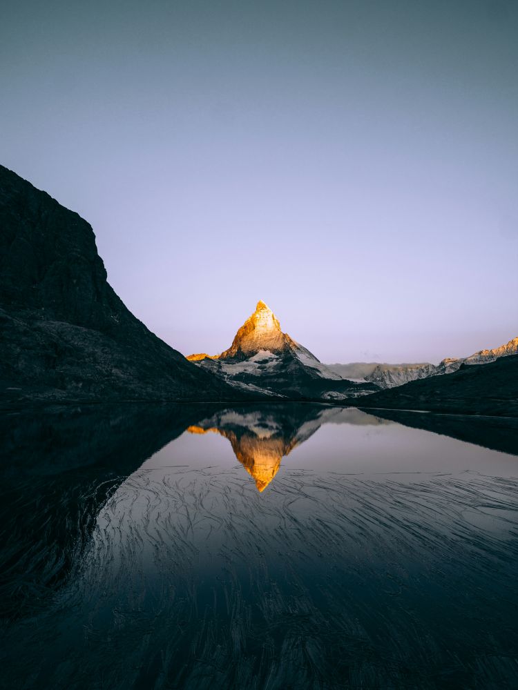 This photo by Robert Richarz shows the snow-capped Matterhorn mountain peak illuminated by sunrise, reflected in a calm alpine lake (Riffelsee) surrounded by dark rocky slopes and clear sky in the Swiss Alps landscape.