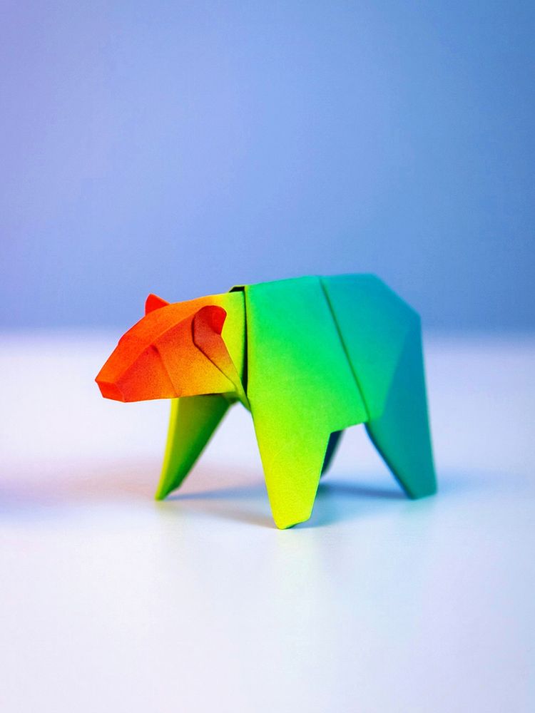 This photo by Florent Fabre shows a colorful origami bear made from gradient paper with orange, yellow, and green hues, displayed on a white surface with a blue background.